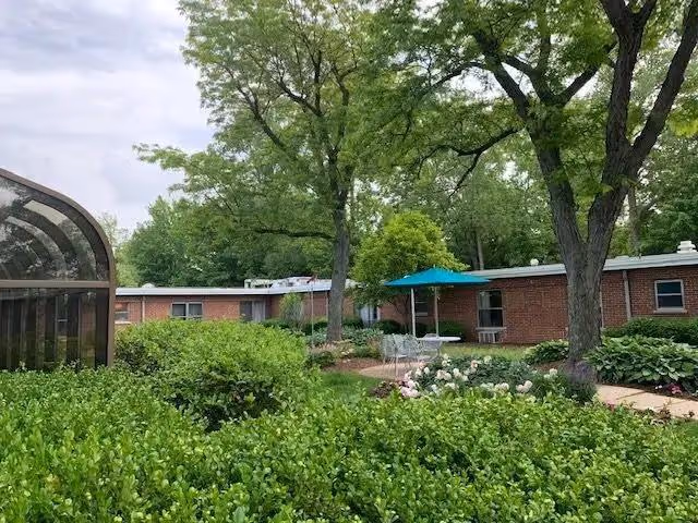 Outdoor garden area at Delmar Gardens of Creve Coeur featuring lush green bushes, trees, a brick building in the background, a patio with a table and chairs under a blue umbrella, and a glass-enclosed structure on the left side.