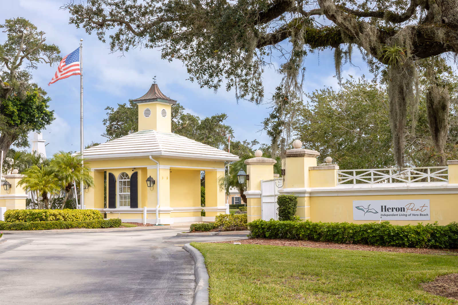 Entrance gate of Heron Point Independent Living of Vero Beach featuring a yellow guardhouse with a white roof, an American flag on a flagpole, and surrounding greenery including trees and bushes.