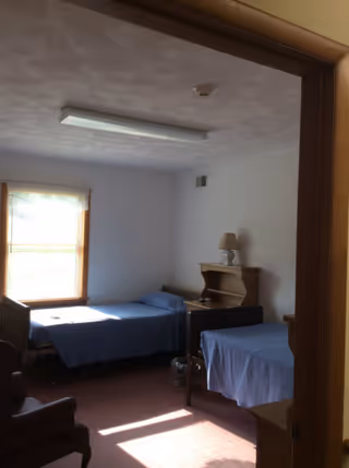A small bedroom with two single beds covered in blue bedspreads, a wooden nightstand with a lamp between the beds, a window with blinds letting in natural light, and a wooden chair partially visible in the foreground.
