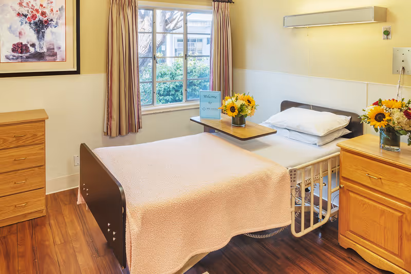 A neatly made single hospital-style bed with a light pink blanket and white pillow in a bright room. There is a wooden bedside table with a vase of sunflowers and another wooden dresser. A window with curtains lets in natural light, and a framed floral painting hangs on the wall.
