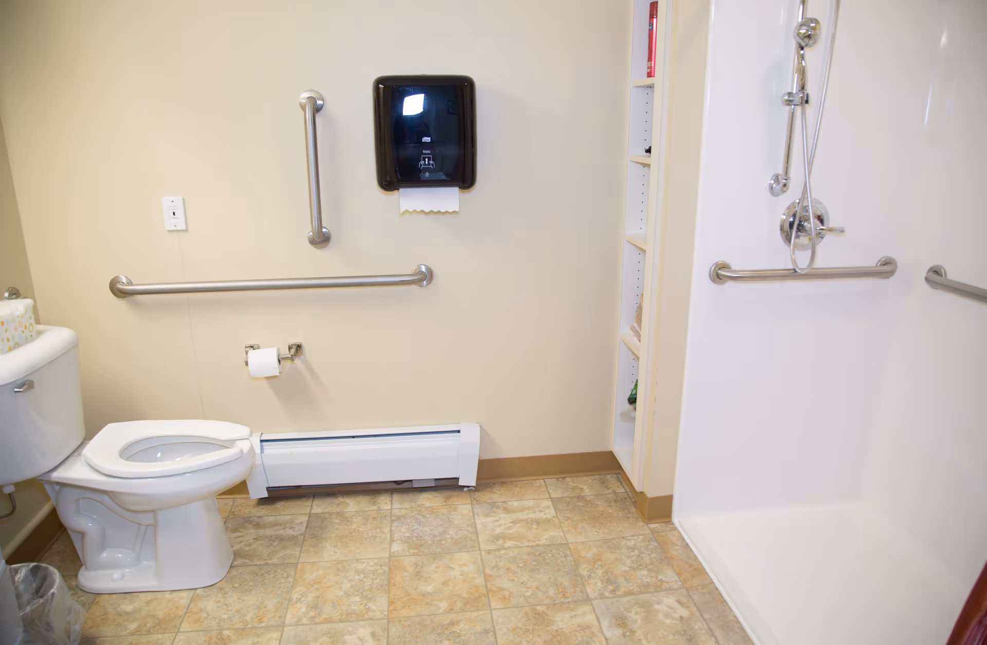 A clean and accessible bathroom with a white toilet, grab bars on the walls, a paper towel dispenser, and a walk-in shower with a handheld showerhead and additional grab bars. The floor is tiled with a beige and brown pattern.