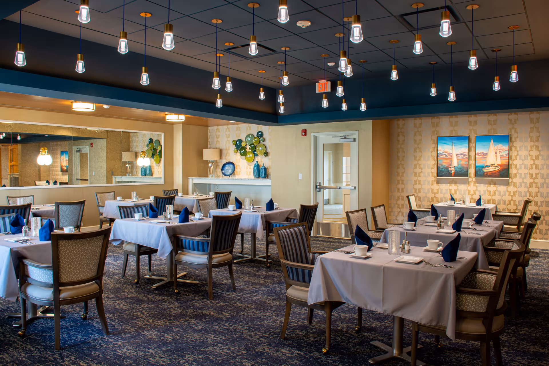 Upscale dining room with multiple tables set with white tablecloths and navy napkins, wooden chairs, pendant lights, and nautical artwork on the wall.
