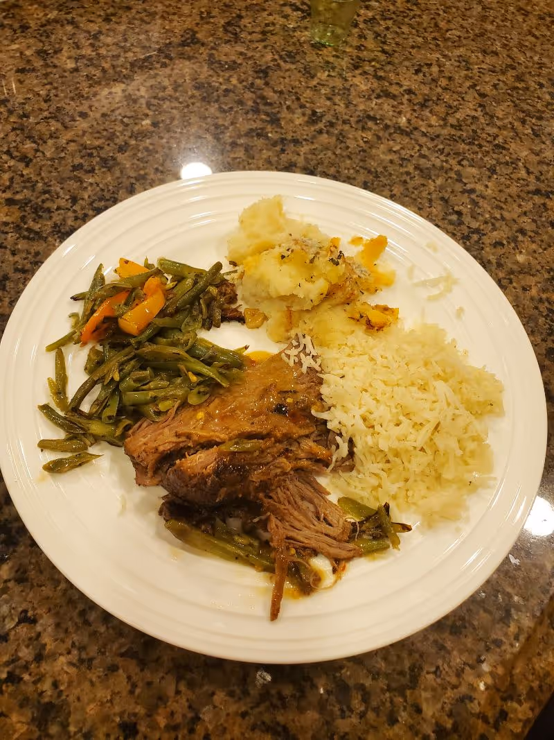 A white plate on a granite countertop with a serving of cooked green beans with carrots, mashed potatoes, shredded beef, and white rice.