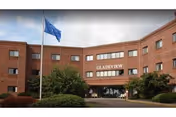 Exterior view of Gladeview Health Care Center, a multi-story brick building with a flagpole flying a blue flag in front, surrounded by bushes and trees.