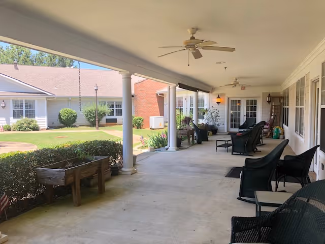 Covered outdoor patio area with ceiling fans, several black wicker chairs, and a small table. The patio overlooks a green lawn with bushes and a building with white siding and a brown roof in the background.