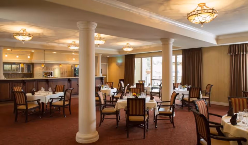 Elegant dining room with round tables set with white tablecloths and chairs, decorative columns, and a serving counter in the background.