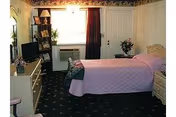 A cozy senior living bedroom with a single bed covered in a pink quilt, a nightstand with a lamp and flowers, a dresser with a TV and framed photos, a window with curtains, and a door leading outside or to another room.