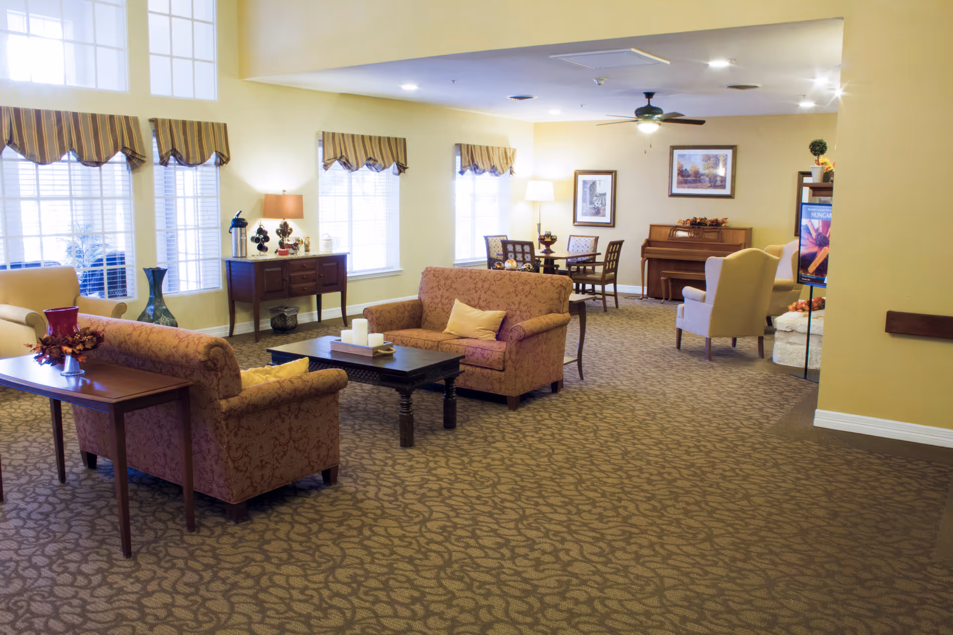 A spacious living room area with patterned carpet, two upholstered sofas with yellow pillows, a dark wooden coffee table with candles, a side table with a red vase, and large windows with striped valances. In the background, there is a dining table with chairs, a piano, framed artwork on the walls, a ceiling fan, and a floor lamp.