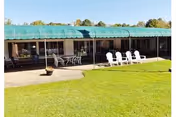 Outdoor patio area of a nursing facility with a green awning covering a walkway. Several white Adirondack chairs and metal tables with chairs are arranged on the concrete patio. The patio overlooks a well-maintained grassy lawn with trees in the background under a clear sky.