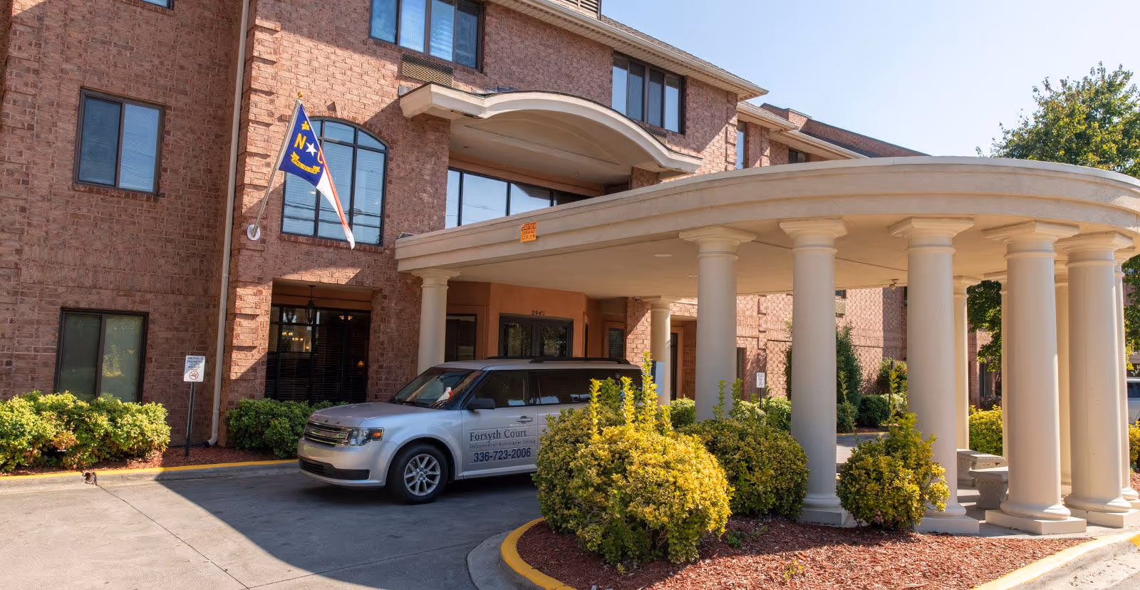 Exterior view of Holiday Forsyth Court building entrance with a covered driveway supported by white columns. A silver vehicle with Forsyth Court signage is parked under the entrance canopy. The building is made of red brick and has several windows. There are green bushes and landscaping around the entrance area, and a North Carolina state flag is displayed on a pole attached to the building.