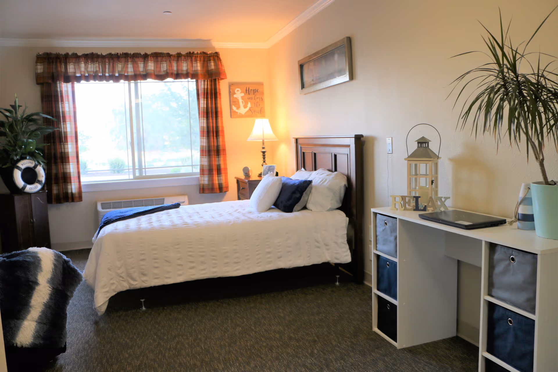 A cozy senior living bedroom with a neatly made bed featuring white bedding and multiple pillows. The room has a large window with plaid curtains letting in natural light. A wooden nightstand with a lamp and a decorative sign that says 'Hope anchors the soul' is beside the bed. Opposite the bed is a white desk with storage bins, a laptop, a lantern, and a plant. Another large plant is in the corner near the window.