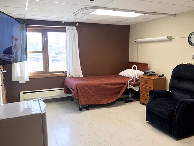 A small bedroom in a senior living facility with a single bed covered in a red blanket, a wooden nightstand with a phone and lamp, a large dark blue recliner chair, a wall-mounted TV showing a black and white image, a window with white curtains, and a clock on the wall.