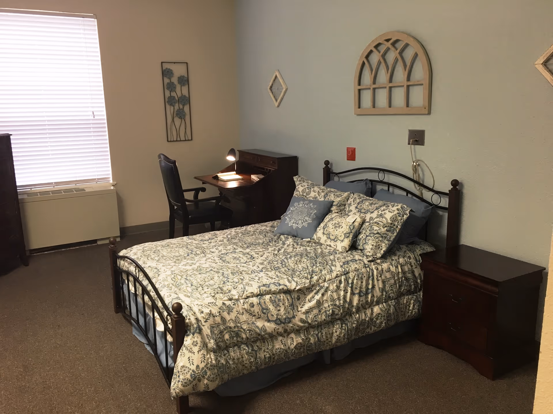 Furnished bedroom with a patterned bed, nightstand, desk and chair against a pale blue wall with decorative accents.