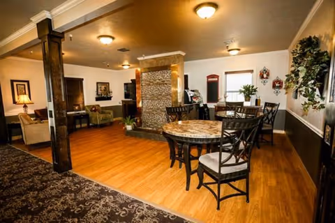 Interior view of a senior living facility common area with wooden flooring, a round table with four chairs, a stone fireplace in the center, and comfortable seating including sofas and armchairs. The room is warmly lit with ceiling lights and decorated with plants and framed artwork.