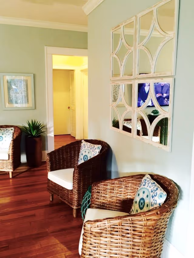 A cozy interior seating area with two wicker chairs featuring white cushions and patterned pillows. The room has light green walls, wooden flooring, a decorative wall mirror with four sections, a framed picture, and a potted plant. An open doorway leads to a hallway with yellow lighting.