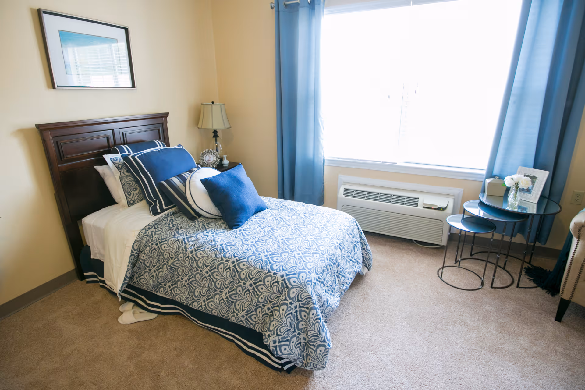 A neatly made single bed with a dark wooden headboard, adorned with multiple blue and white pillows and a patterned blue and white bedspread. Next to the bed is a small nightstand with a lamp and a framed photo. A large window with blue curtains lets in bright natural light. In front of the window is a white air conditioning unit. To the right, there is a set of three nested round glass tables with a small vase of white flowers and a framed picture on top. The room has beige walls and carpeted floor.