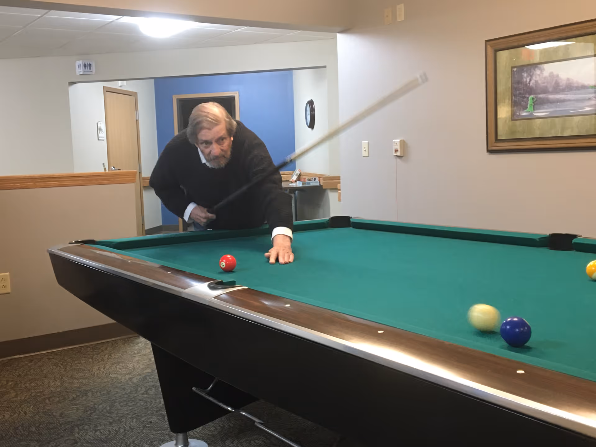 An elderly man playing pool indoors, aiming with a cue stick at a billiard table with several balls on the green felt surface. The room has beige walls, a framed picture on the wall, and a blue accent wall in the background.