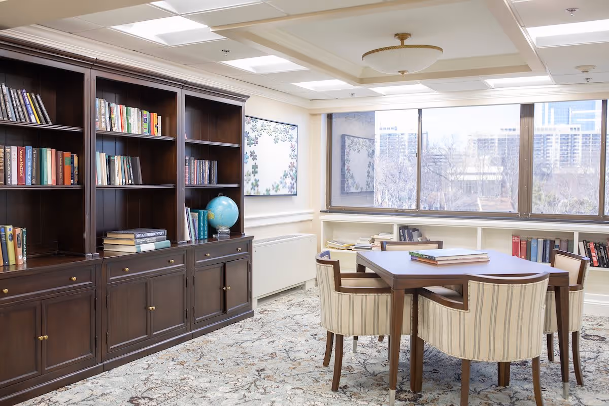 A bright room with large windows showing an outdoor cityscape. Inside, there is a wooden table surrounded by four striped upholstered chairs. Along one wall, there is a large dark wooden bookshelf filled with books and a globe. The floor is covered with a patterned carpet, and there is a framed artwork on the wall.