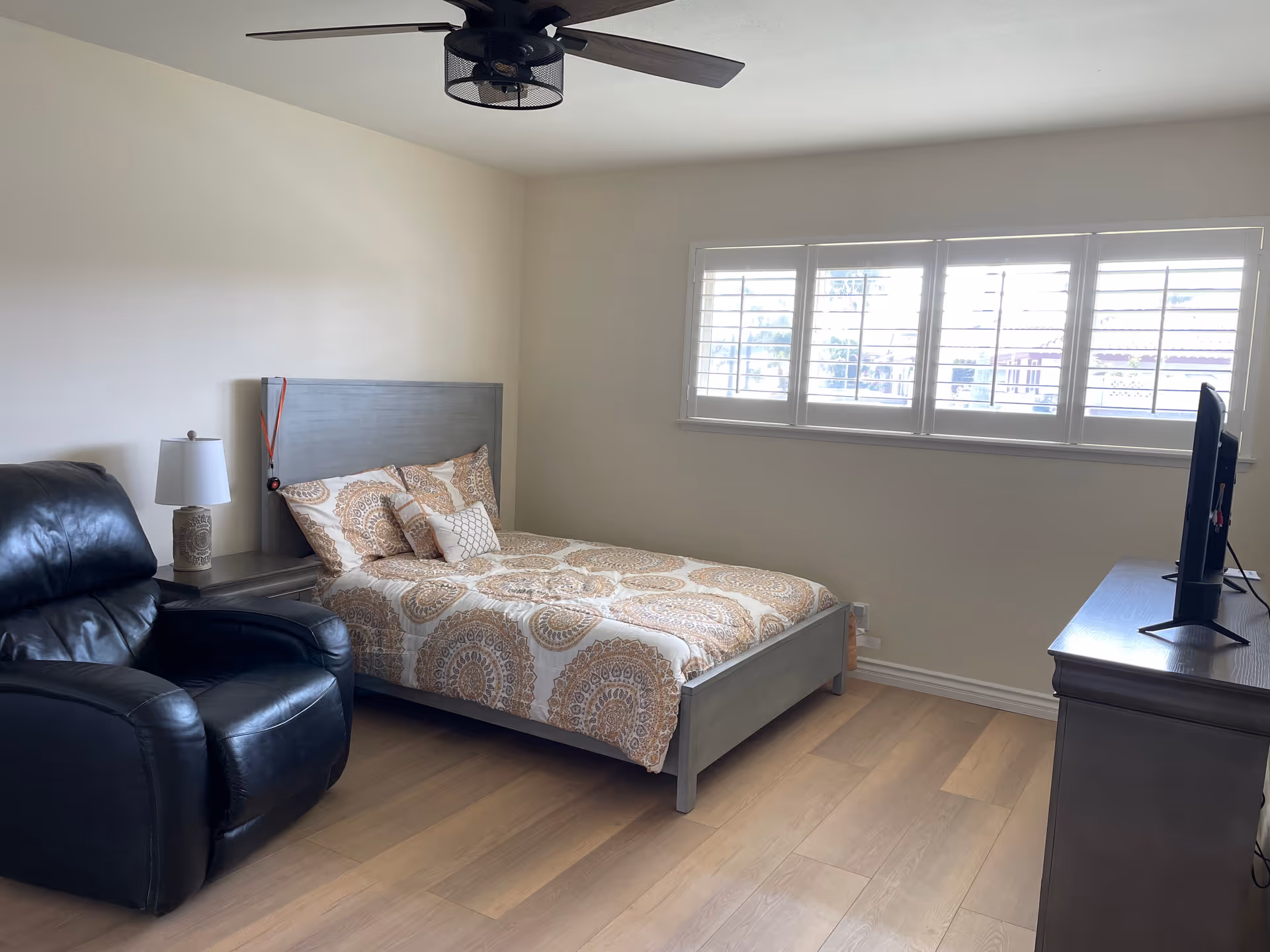 Sunny bedroom with a gray bed and patterned comforter, black leather recliner, dresser with a TV, and shuttered windows.