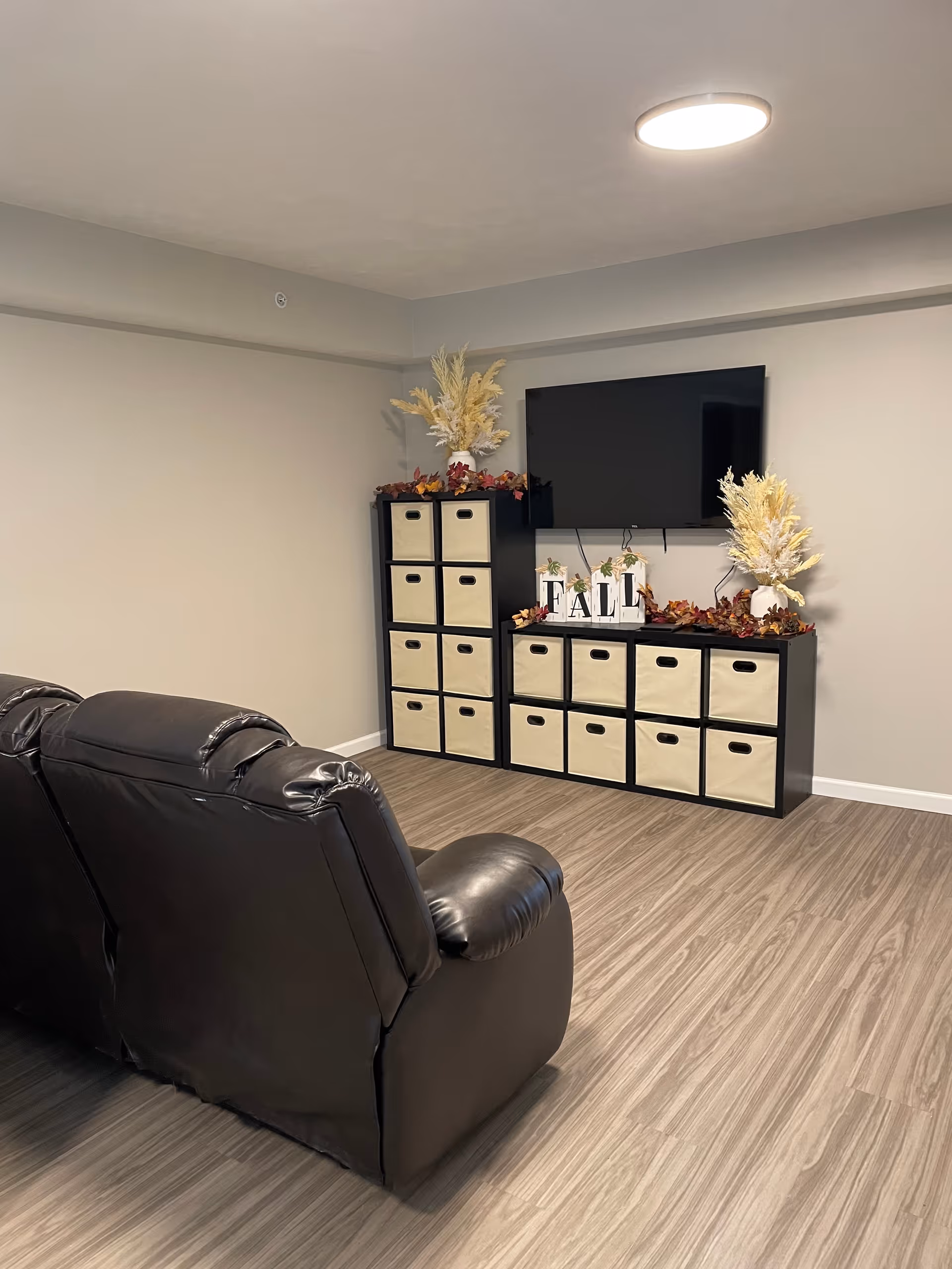 A cozy living room area with a black leather recliner sofa facing a wall-mounted flat screen TV. Below the TV are two black storage units with beige fabric bins. The top of the storage units is decorated with fall-themed decor including artificial plants and blocks spelling out 'FALL'. The room has light-colored walls and wood-look flooring with a round ceiling light.