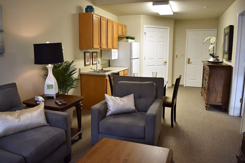 A cozy living area with gray upholstered armchairs and a matching sofa, each with decorative pillows. A wooden side table with a white lamp and a remote control sits beside the sofa. In the background, there is a small kitchen area with wooden cabinets, a white refrigerator, a microwave, and a sink. A dining table with chairs is positioned near two white doors. A wooden dresser with a white orchid plant and a framed picture hangs on the wall. The room has beige walls and carpeted flooring.