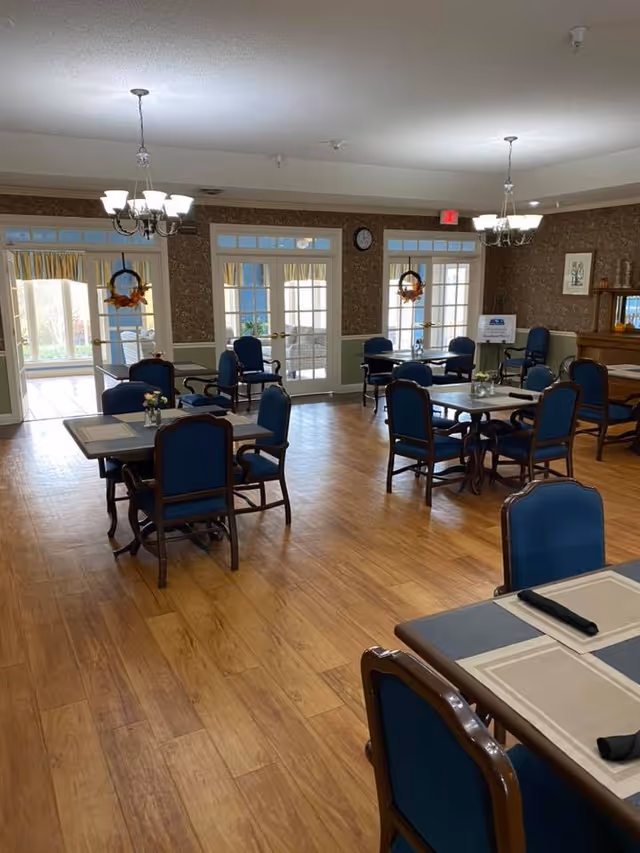 Spacious dining room with several square tables and blue upholstered chairs on a wood floor, chandeliers, and French doors with wreaths.