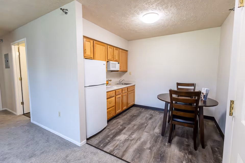Small kitchen area with wooden cabinets, a white refrigerator, a microwave, and a sink. Adjacent to the kitchen is a dining area with a round wooden table and three chairs. The floor in the kitchen and dining area is wood-patterned, while the adjacent hallway has carpeted flooring.
