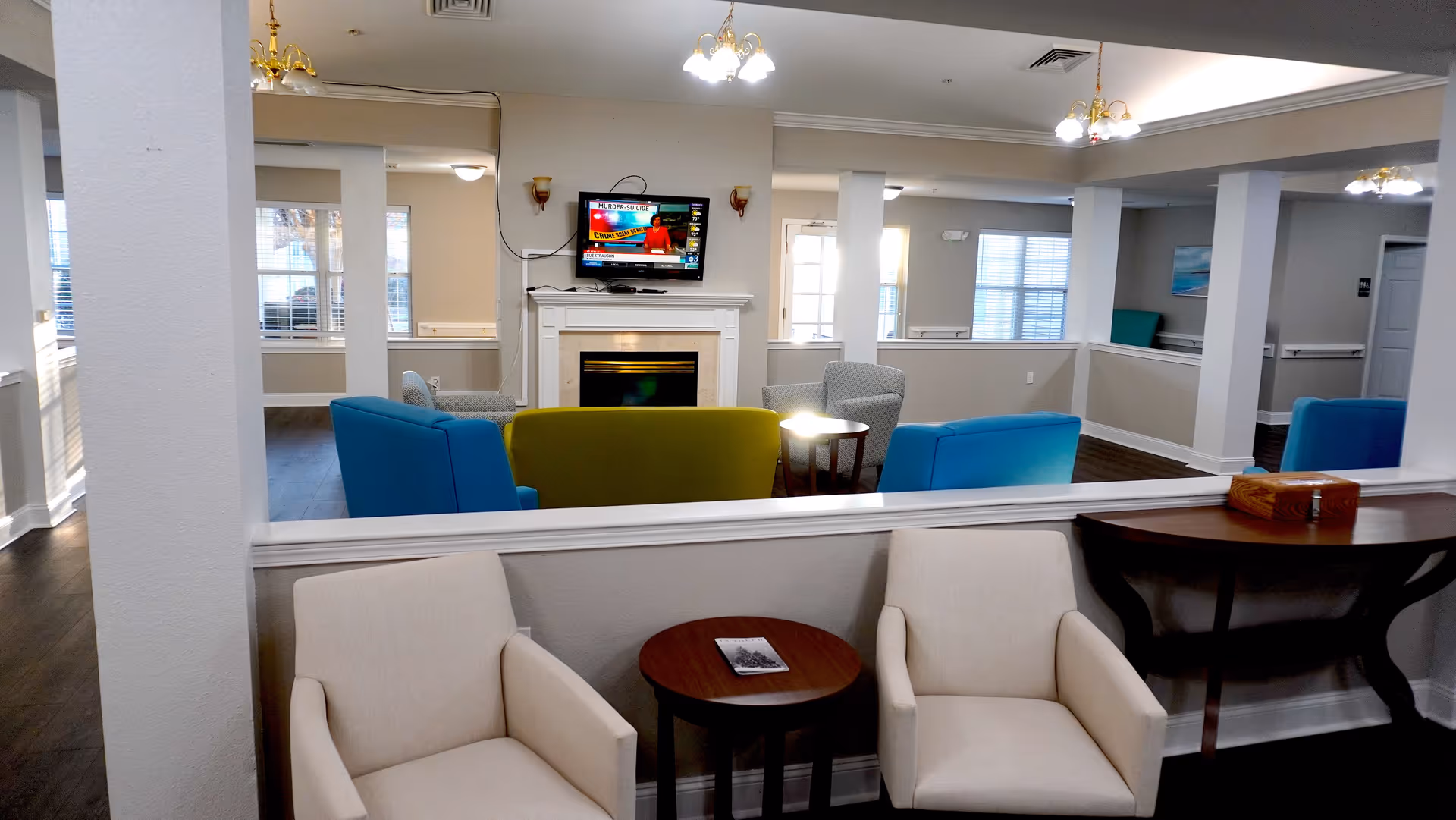 A cozy common area in a senior living facility with two beige armchairs and a small round wooden table in the foreground. Beyond a half wall, there are colorful chairs including blue and green ones arranged around a fireplace with a mounted flat-screen TV above it. The room has multiple windows letting in natural light and several ceiling light fixtures.