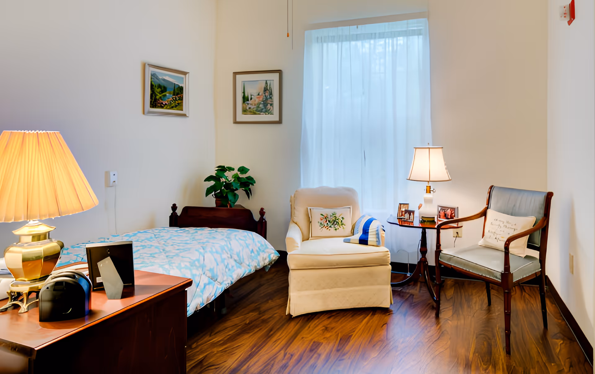 A sunlit senior living bedroom with a single bed, an upholstered armchair and wooden chair beside a small table with lamps on a wood floor.