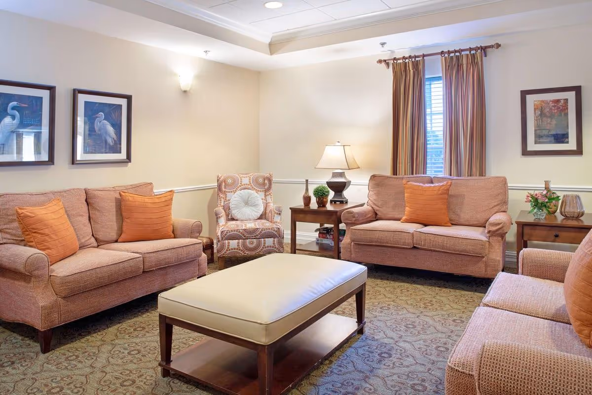A cozy living room with three peach-colored sofas, each with orange throw pillows, and a patterned armchair with a white round pillow. There is a large rectangular ottoman in the center with a wooden base and cream-colored cushion. The room has beige walls adorned with framed artwork, a window with striped curtains, two wooden side tables with a lamp, plants, and decorative items, and a patterned carpet on the floor.