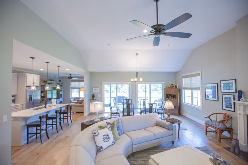 Bright and spacious open-concept living area with a curved light gray sofa adorned with decorative pillows, a wooden side table with a lamp, and a ceiling fan above. The room features large windows and sliding glass doors that let in natural light. Adjacent to the living area is a dining space with a glass-top table and chairs, and a kitchen with a long island and bar stools. The walls are painted light gray, and the flooring is light wood.