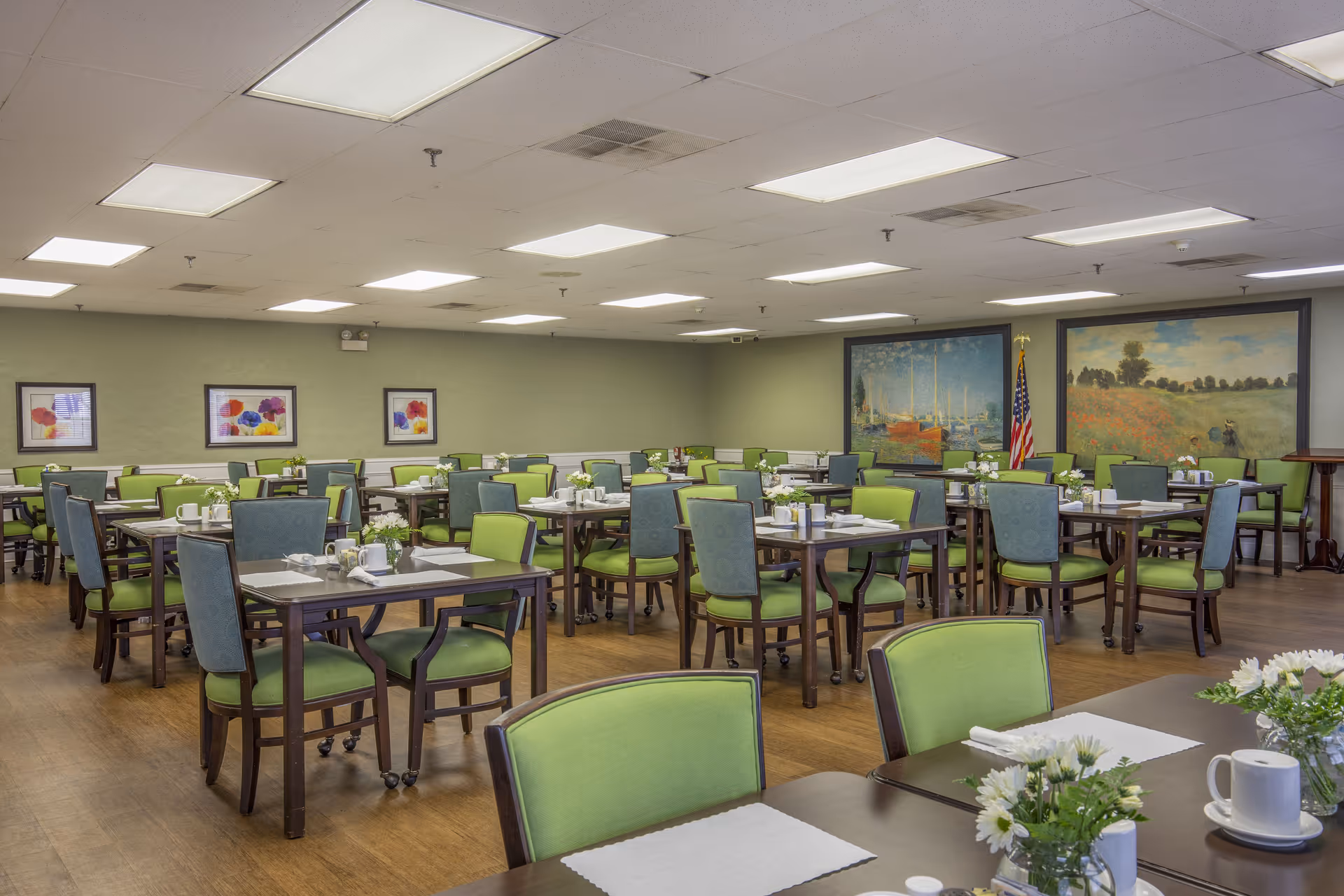 Spacious dining room with multiple tables and green upholstered chairs set for service and paintings on the walls.