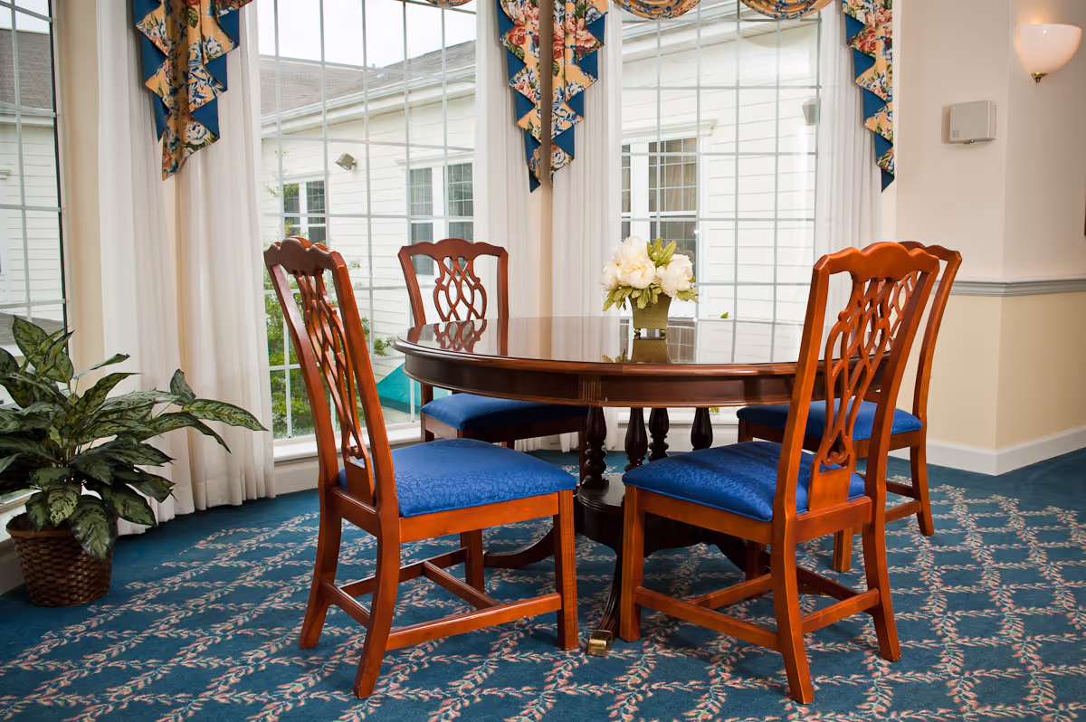 A round wooden dining table with four wooden chairs featuring blue cushioned seats, placed on a patterned blue carpet. The room has large windows with white curtains and floral valances, and a potted plant is visible to the left of the table. A small flower arrangement is centered on the table.
