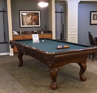 A pool table with a green felt surface and balls arranged on it in a room with beige carpet and gray walls. There is a framed picture of a horse on the wall above a wooden cabinet with decorative items. A white sign is placed on the pool table. The room has columns and an archway leading to another area.