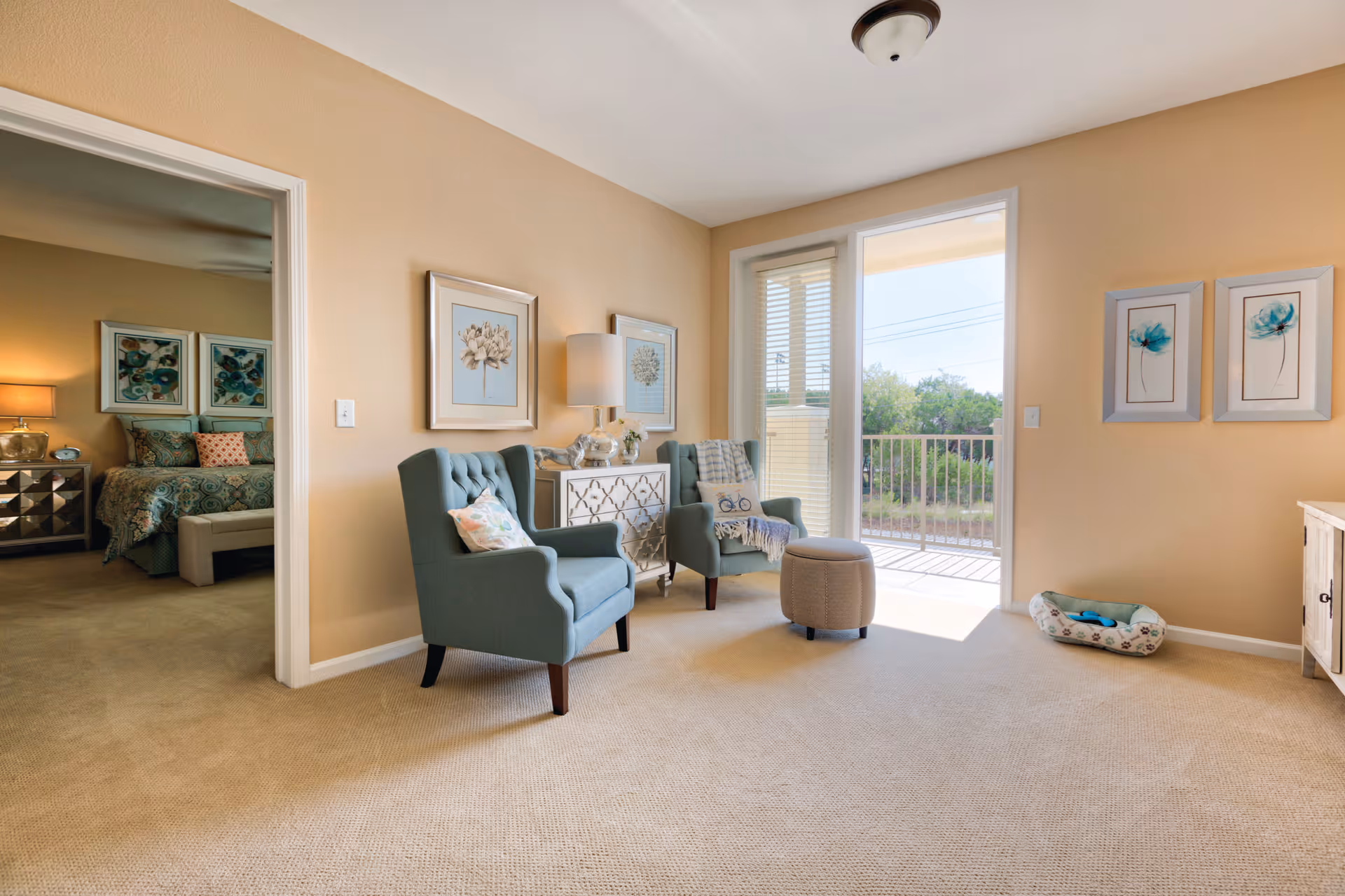 A bright living room with beige walls and carpet, featuring two blue armchairs with decorative pillows, a small ottoman, and a white cabinet with floral artwork above it. There is a sliding glass door leading to a balcony with a view of greenery outside. To the left, an open doorway reveals a bedroom with a bed, nightstand, and lamps.