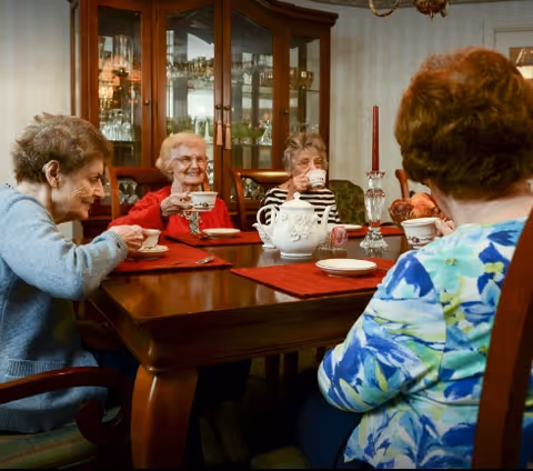 Four elderly women sitting around a wooden dining table enjoying tea together in a cozy dining room with a china cabinet in the background.