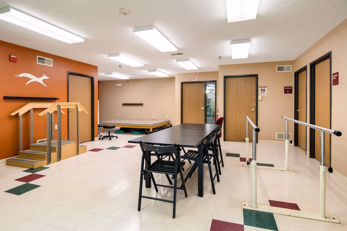 Activity and therapy room with a long table and folding chairs, parallel walking bars, exercise stairs, and a treatment table.