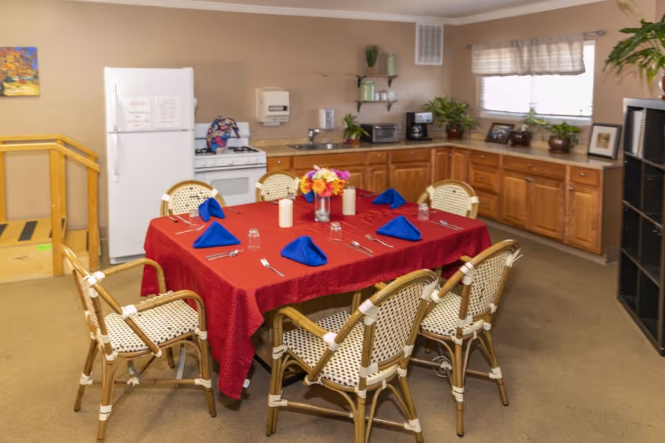 A dining area with a rectangular table covered with a red tablecloth, set with blue folded napkins, glasses, and silverware. The table is surrounded by eight wicker chairs with white and brown woven seats and backs. In the background, there is a kitchen area with wooden cabinets, a white refrigerator, a stove, a microwave, a coffee maker, and several potted plants on the countertop. A window with a valance lets in natural light, and there is a small staircase with a wooden handrail on the left side.