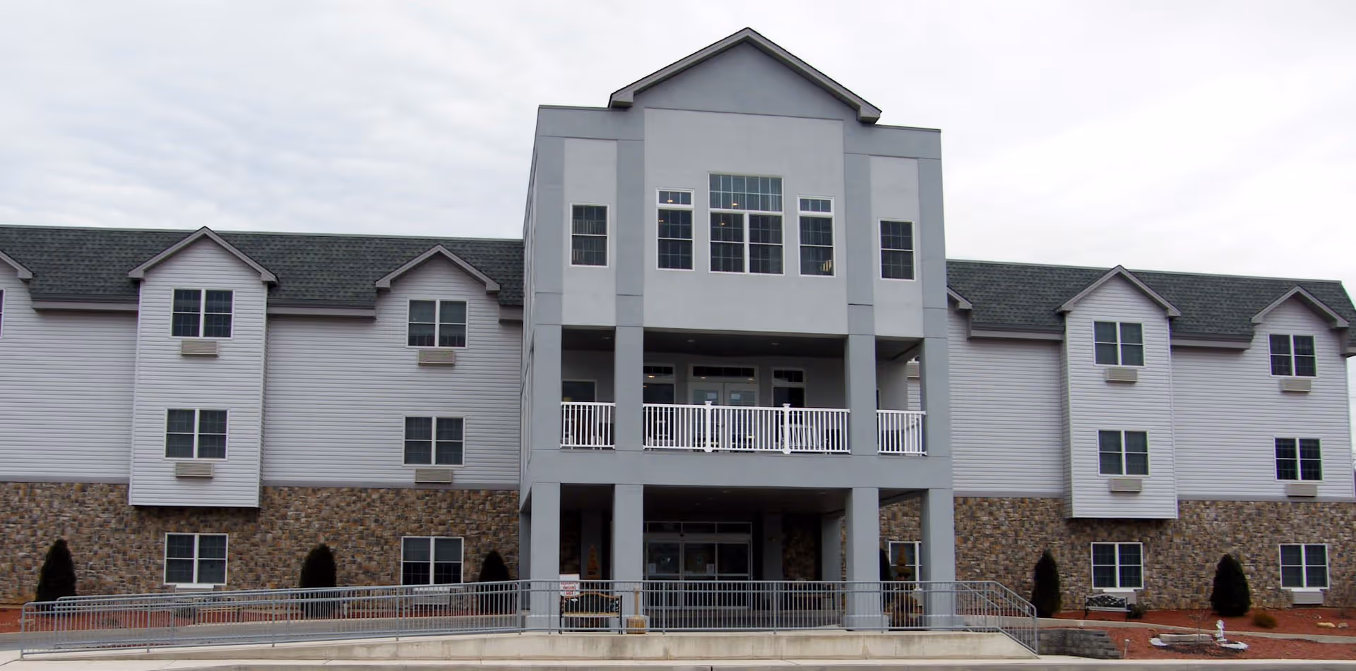 Front exterior view of The Palmerton facility, a three-story building with a combination of stone and light-colored siding, multiple windows, and a central entrance with a balcony above it. The building has a pitched roof and a ramp leading to the entrance.