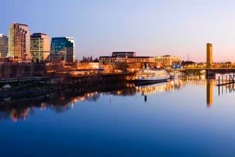 Calm river reflects a city skyline with boats and illuminated buildings at dusk.