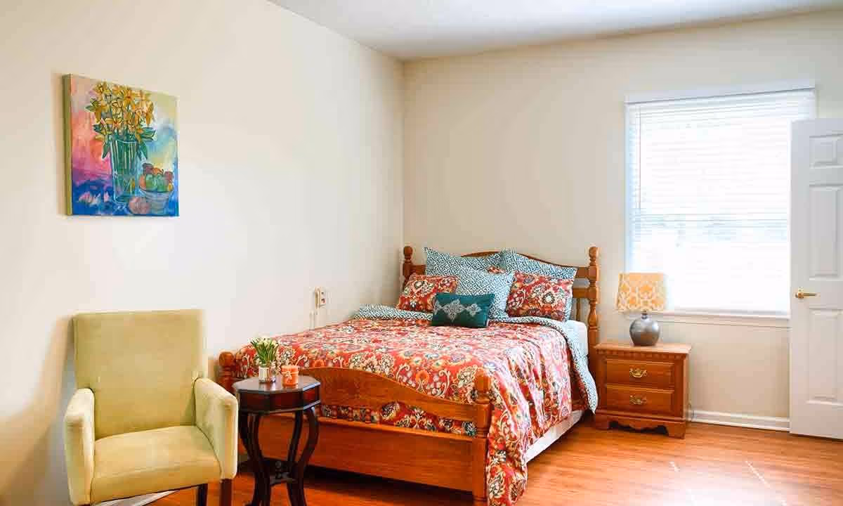A cozy bedroom with a wooden bed featuring a colorful floral bedspread and multiple pillows. Next to the bed is a wooden nightstand with a lamp, and a window with blinds letting in natural light. A light green upholstered armchair and a small round wooden side table with a plant and a cup are placed near the bed. A colorful painting of flowers hangs on the wall above the chair.