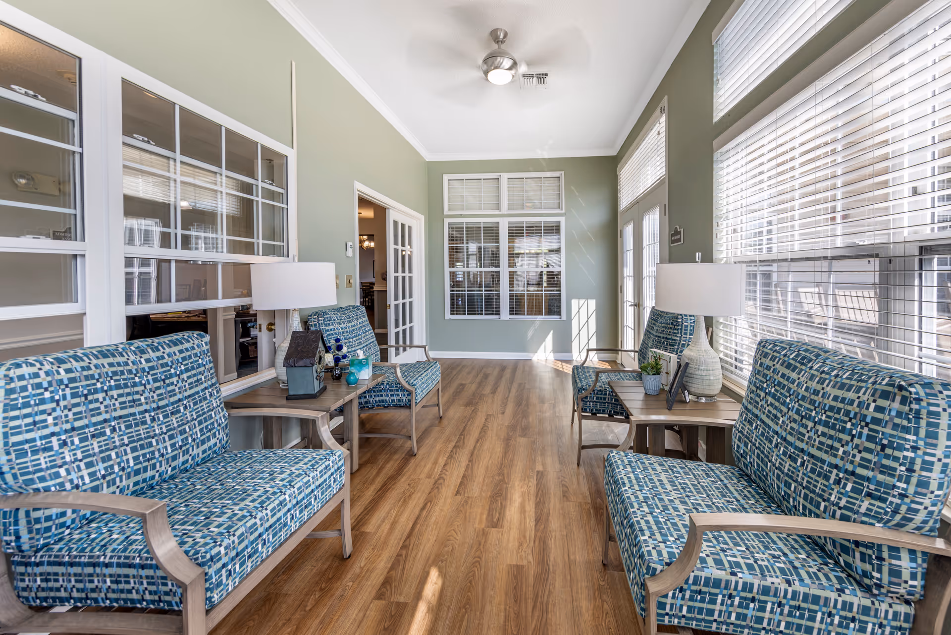 A bright and airy sitting area with four blue patterned cushioned chairs arranged in pairs facing each other along the walls. Between each pair of chairs are wooden side tables with white lamps and decorative items. The room has light green walls, large windows with white blinds, and wood flooring. A ceiling fan with a light is mounted on the white ceiling.