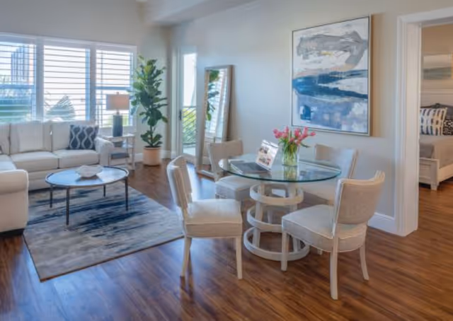 A bright and modern living room and dining area with wooden flooring. The living room features a white sofa with cushions, a round coffee table on a blue and gray rug, and a large window with white blinds. The dining area has a round glass table with four upholstered chairs and a vase of pink flowers on top. A tall mirror leans against the wall next to a potted plant, and a large abstract painting hangs on the wall. A doorway leads to a bedroom with a bed and pillows visible.