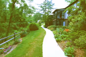 A curved concrete walkway passes through a landscaped garden beside a two-story building surrounded by trees and shrubs.