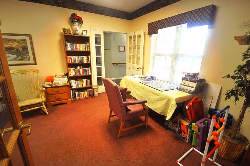 A cozy room with a red carpet and yellow walls featuring a bookshelf filled with books, a wooden rocking chair, a table covered with a yellow cloth holding a puzzle and stacks of board games, two upholstered chairs, a large window with a patterned valance, and a potted plant in the corner.
