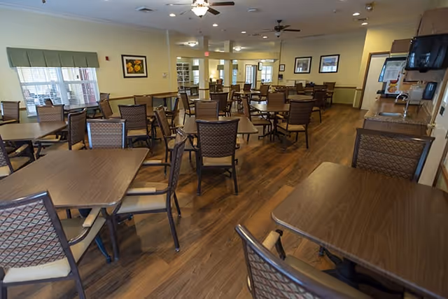 A spacious dining room with multiple wooden tables and cushioned chairs arranged neatly on a wooden floor. The room has large windows with blinds, framed artwork on the walls, ceiling fans, and soft lighting. There is a counter area on the right side with a microwave and some informational materials.