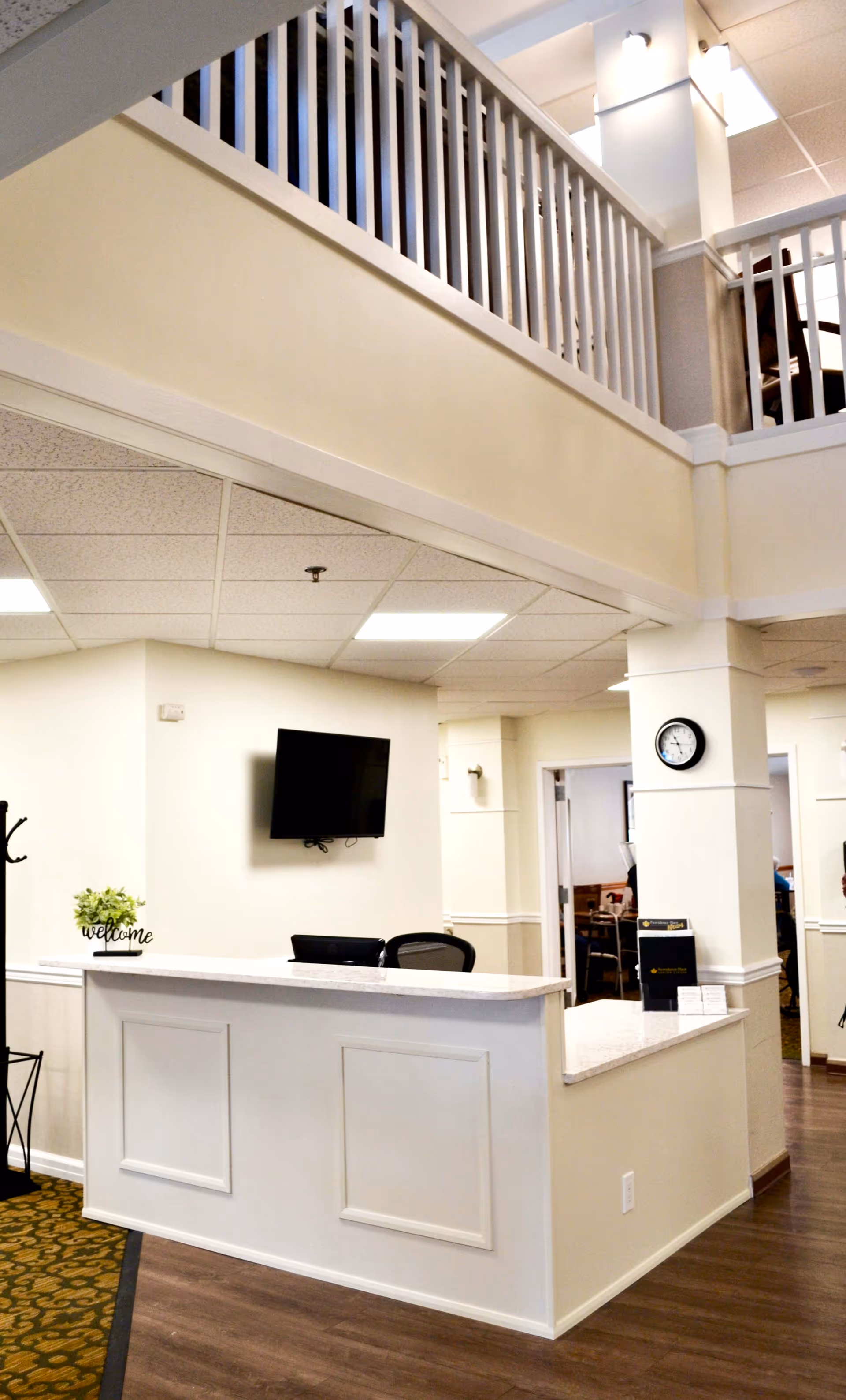 Reception area inside Providence Place Senior Living of Pine Grove with a white front desk, a black office chair, a wall-mounted TV, a small plant with a welcome sign, a clock on the wall, and an upper balcony railing visible above.