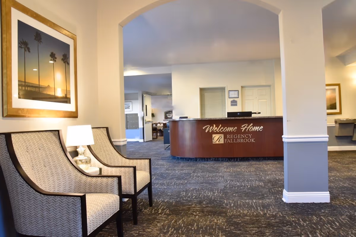 Reception area of Regency Fallbrook facility with a wooden front desk that has a 'Welcome Home' sign. Two patterned armchairs and a lamp are placed on the left side under a framed picture of palm trees at sunset. The area has carpeted flooring and light-colored walls with an archway leading to the reception desk.