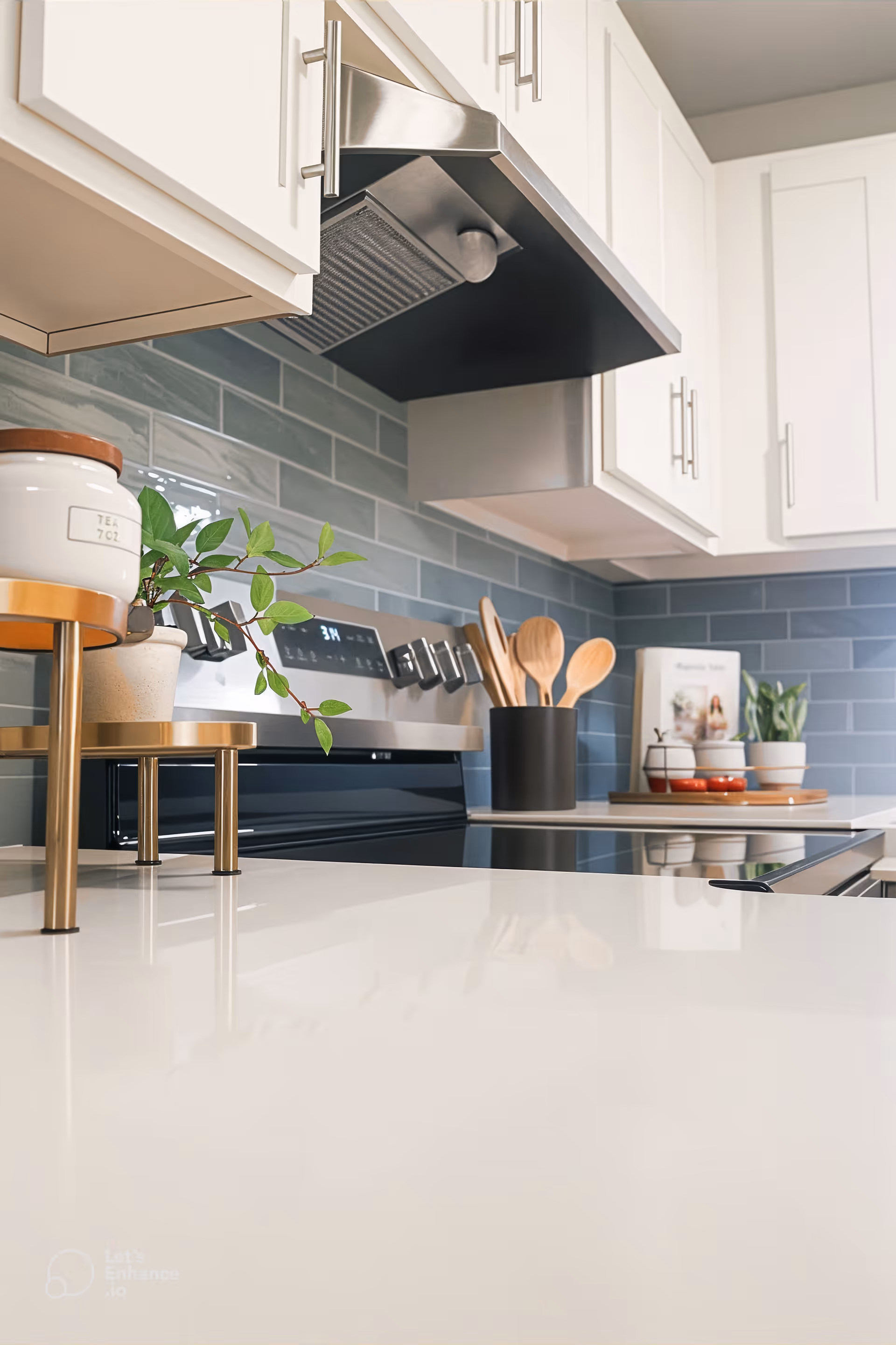 Modern kitchen with white cabinets, a stainless steel stove and range hood, blue subway tile backsplash, wooden cooking utensils in a black holder, potted plants, and a clean white countertop.