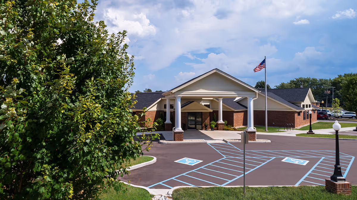 Exterior view of Rawlins House Family-first Senior Living facility showing the main entrance with a covered drop-off area supported by white columns, a parking lot with handicap spaces marked in blue, a flagpole with an American flag, and surrounding greenery under a partly cloudy sky.